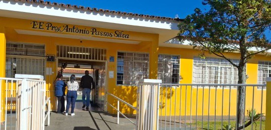 Foto da fachada de uma escola, na cor amarela. Três pessoas estão de pé junto à porta de entrada.