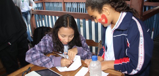 A girl stands looking at another who is sitting, signing a paper.