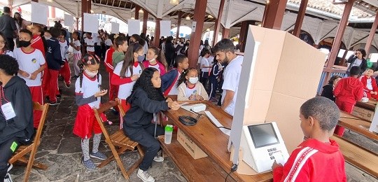 Em primeiro plano, menino vota na urna eletrônica. Ao fundo, várias crianças com uniforme de esc...