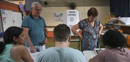 Foto mostra um idoso e uma idosa se identificando junto aos mesários de uma seção eleitoral