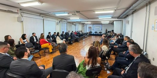 Foto mostra dezenas de pessoas sentadas em uma sala, em cadeiras dispostas em formato de U. Ao f...