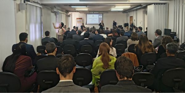 Foto mostra dezenas de pessoas sentadas em uma sala. Ao fundo, o desembargador Paulo Tamburini e...