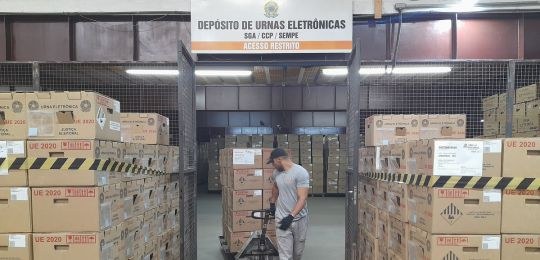 Homem puxa um carrinho com várias caixas. Ele está saindo de um depósito que tem uma placa "Depó...