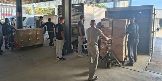 Alguns homens empurram pallets com caixas de urnas eletrônicas, perto de dois caminhões.