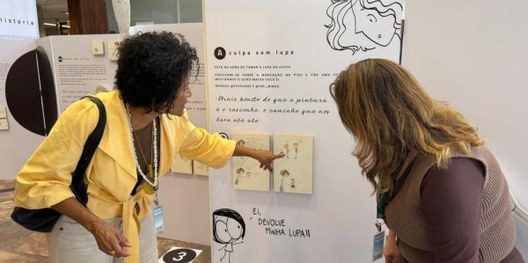 Foto mostra duas mulheres interagindo com elementos nos painéis de uma exposição.