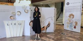 Foto mostra a artista Lu Simão em frente a um painel de exposição.