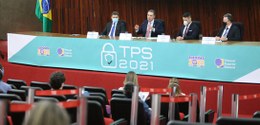 Em uma mesa grande com a inscrição TPS 2021, estão sentadas quatro pessoas.  Ao centro está o Mi...