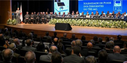 Foto mostra um teatro lotado. Ao fundo, diversas pessoas sentadas junto a uma mesa sobre o palco.