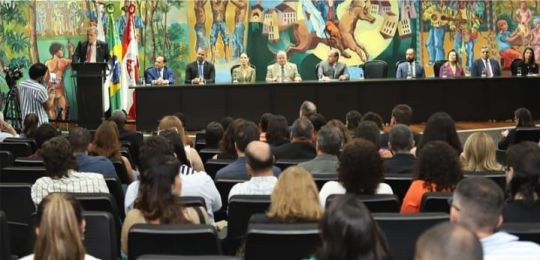 Em primeiro plano, pessoas sentadas na plateia de um auditório, de costas para a câmera. Ao fund...