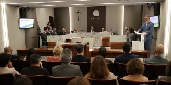 Foto mostra autoridades sentadas junto a uma mesa. Em primeiro plano, parte da plateia na Sala d...