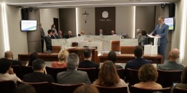 Foto mostra autoridades sentadas junto a uma mesa. Em primeiro plano, parte da plateia na Sala d...