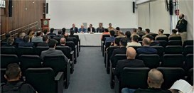 Foto mostra várias pessoas sentadas na plateia de um auditório. Ao fundo, cinco homens sentados ...