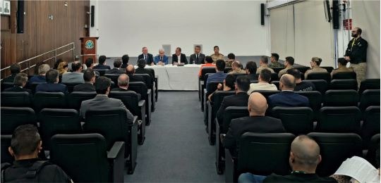 Foto mostra várias pessoas sentadas na plateia de um auditório. Ao fundo, cinco homens sentados ...