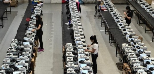 Foto tirada do alto mostra três mulheres junto a bancadas com dezenas de urnas eletrônicas