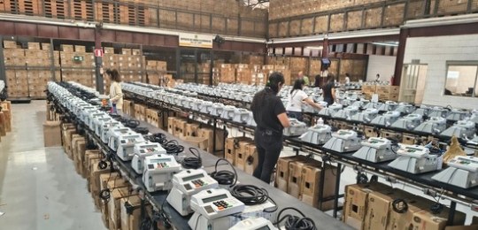 Foto tirada do alto mostra três mulheres junto a bancadas com dezenas de urnas eletrônicas
