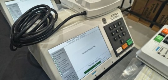 Foto tirada do alto mostra três mulheres junto a bancadas com dezenas de urnas eletrônicas