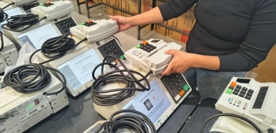 Foto tirada do alto mostra três mulheres junto a bancadas com dezenas de urnas eletrônicas