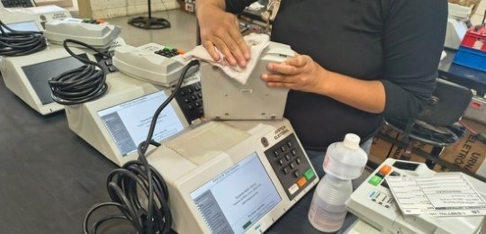 Foto tirada do alto mostra três mulheres junto a bancadas com dezenas de urnas eletrônicas