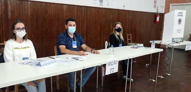 A foto mostra três mesários sentados e ao fundo aparece uma cabine eleitoral