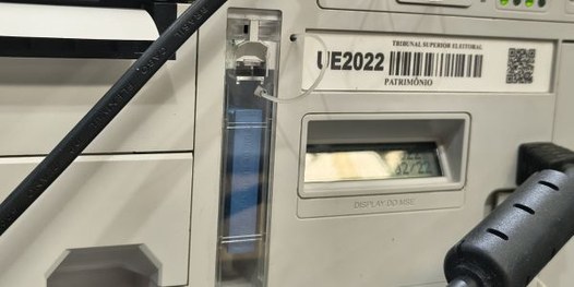 Foto mostra dezenas de urnas eletrônicas sobre bancadas, dentro de um galpão. Algumas pessoas es...