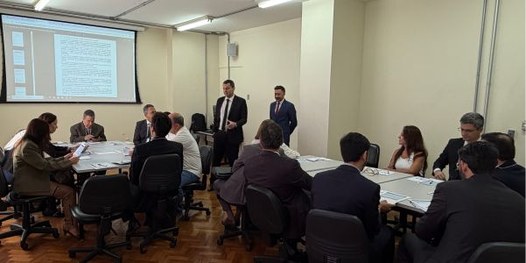 Foto mostra dezenas de pessoas sentadas em uma sala, em cadeiras dispostas em formato de U. Ao f...