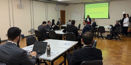 Foto mostra dezenas de pessoas sentadas em uma sala, em cadeiras dispostas em formato de U. Ao f...