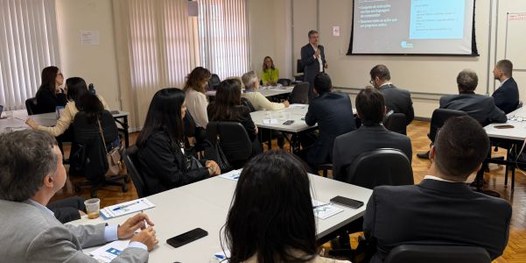 Foto mostra dezenas de pessoas sentadas em uma sala, em cadeiras dispostas em formato de U. Ao f...