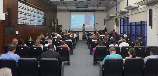 Foto de um auditório lotado. Ao fundo, a diretora-geral do TRE, Glória Araújo, está de pé no palco.