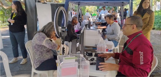 Ouvidoria Itinerante 2025 Foto mostra mesas com guichês de atendimento ao eleitor. Dois servidores atendem eleitores. Ao r...