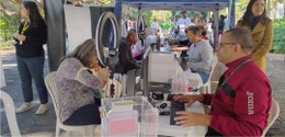 Foto mostra mesas com guichês de atendimento ao eleitor. Dois servidores atendem eleitores. Ao r...