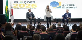 Foto da ministra Cármen Lúcia de pé durante palestra. Em primeiro plano, de costas, estudantes s...