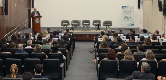 Foto mostra parte do auditório do TRE-MG, com pessoas sentadas na plateia, olhando para o palco....