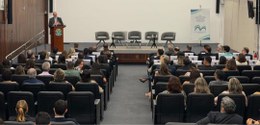 Foto mostra parte do auditório do TRE-MG, com pessoas sentadas na plateia, olhando para o palco....