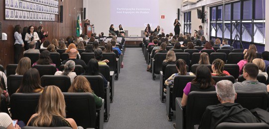 Foto de auditório lotado. Ao fundo, quatro mulheres sentadas em cadeiras sobre um palco.