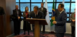 Desembargador Júlio César Lorens, de pé, assina um documento. Ele é observado pelo desembargador...