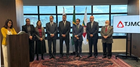 Duas mulheres e cinco homens, de pé, posam para a foto. No canto esquerdo, há mais uma mulher, j...