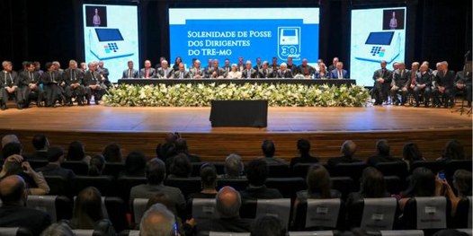 Foto mostra um teatro lotado. Ao fundo, diversas pessoas sentadas junto a uma mesa sobre o palco.