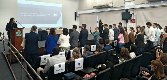 Foto mostra várias pessoas de pé na plateia de um auditório, com a mão direita levantada. No can...