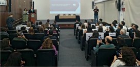Foto mostra dezenas de pessoas na plateia de um auditório. Ao fundo, de pé, está o desembargador...