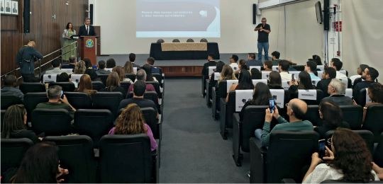 Foto mostra dezenas de pessoas na plateia de um auditório. Ao fundo, de pé, está o desembargador...