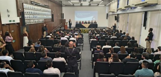 Foto mostra a plateia de um auditório. Ao fundo, algumas pessoas sentadas junto a uma mesa em ci...