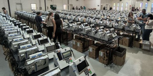 Foto mostra dezenas de urnas eletrônicas sobre bancadas, dentro de um galpão. Algumas pessoas es...