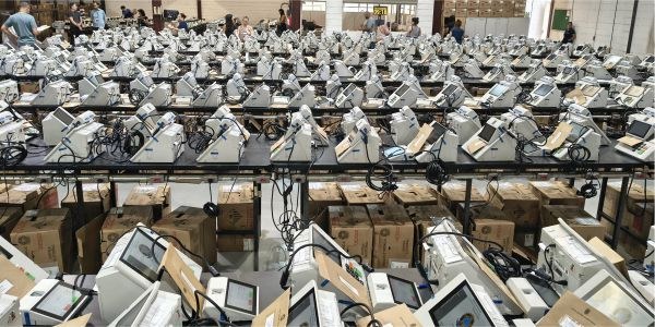 Foto mostra dezenas de urnas eletrônicas sobre bancadas, dentro de um galpão.