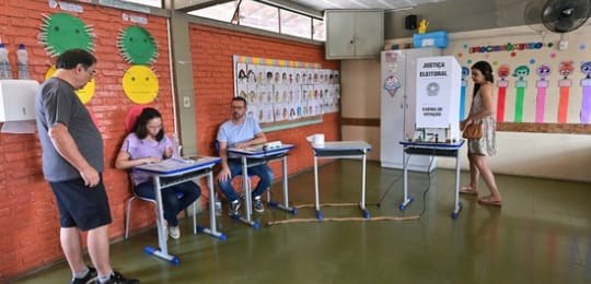 primeiro turno MG 2024 Um homem de pé espera junto a mesas onde estão sentados dois mesários. Ao fundo, há uma eleitora...