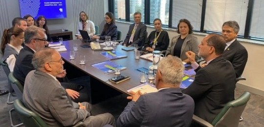 reunião Comitê Bio TRE-MG Homens e mulheres sentados ao redor de uma mesa de madeira oval. No canto direito, o desembargad...