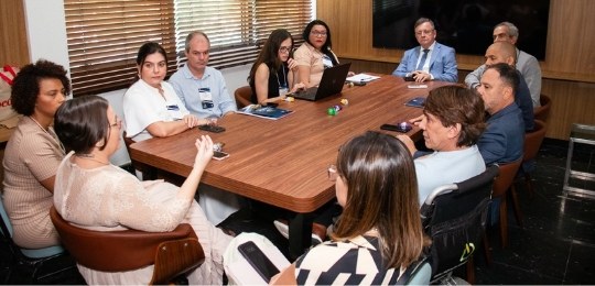 Fota mostra homens e mulheres sentados ao redor de uma grande mesa em formato de U.