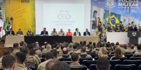 Foto de auditório. No primeiro plano, há pessoas de costas e ao fundo, uma mesa com autoridades ...