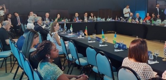 Fota mostra homens e mulheres sentados ao redor de uma grande mesa em formato de U.