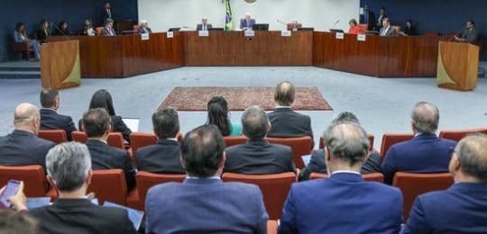 Foto mostra ministro Edson Fachin, ao fundo, sentado junto a uma mesa com outras autoridades. Em...