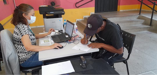 Servidora do TRE-MG observa um homem de boné assinar um documento.